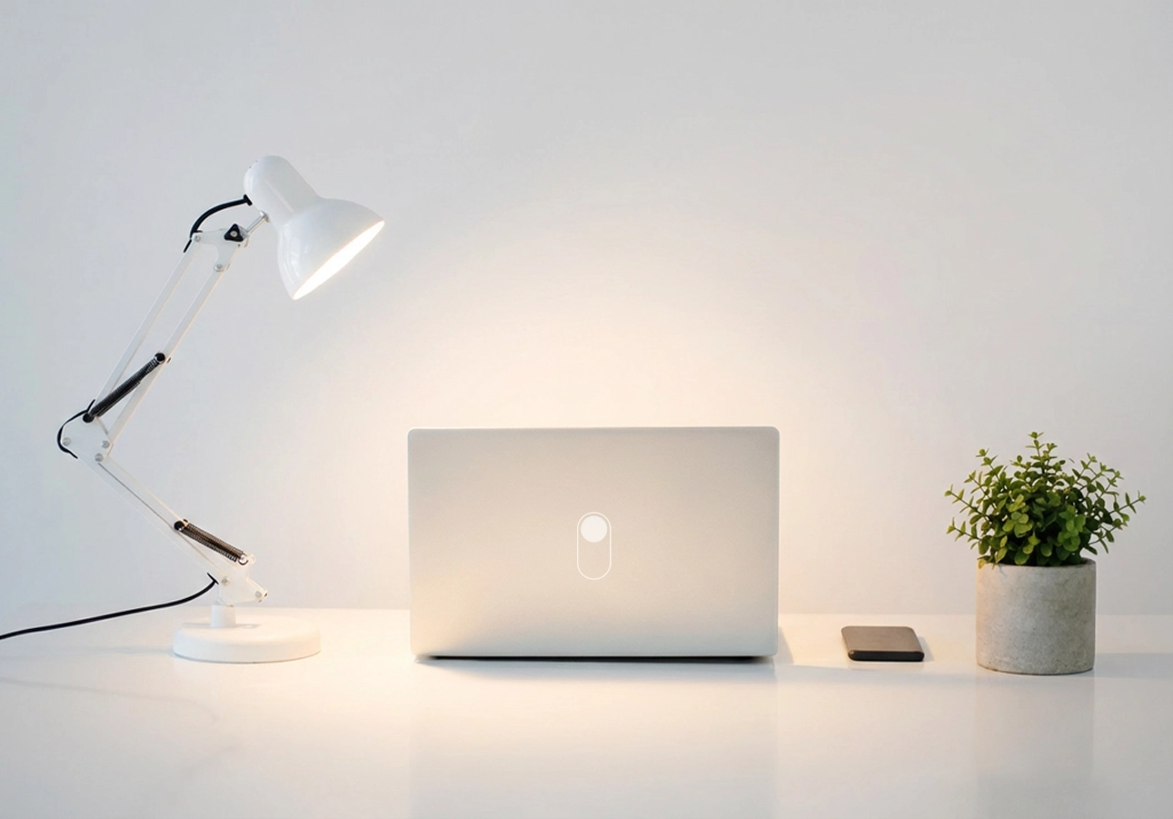 Minimalistischer weißer Schreibtisch mit Laptop, Schreibtischlampe, kleiner Topfpflanze und Smartphone vor einer hellen Wand.